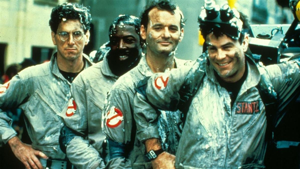 Four Ghostbusters in gray jumpsuits smiling and covered in marshmallow-white goo in a film still