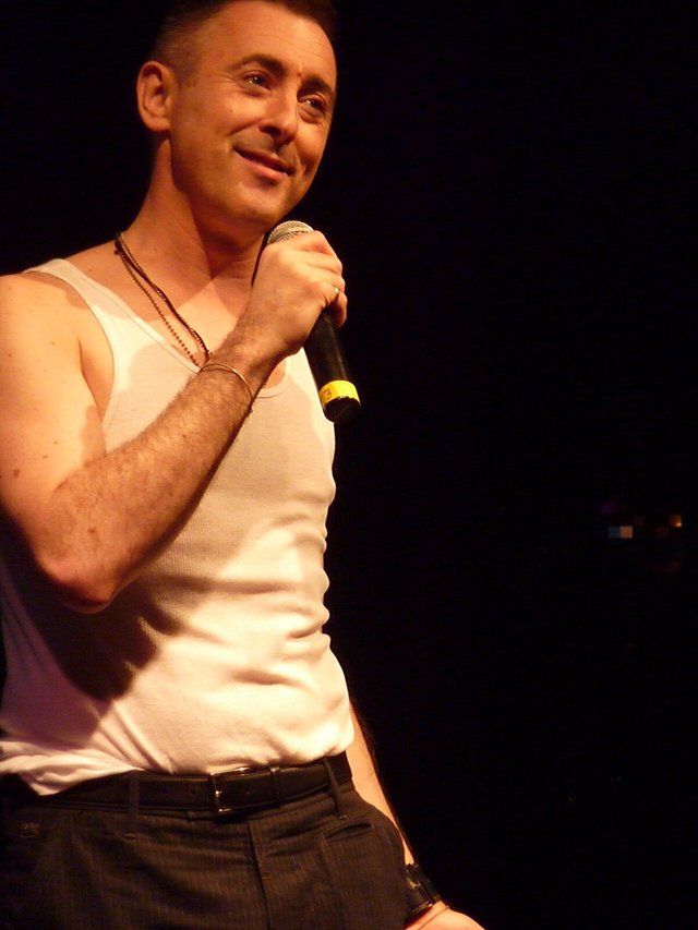 Alan Cumming smiling and holding a microphone on a dark stage