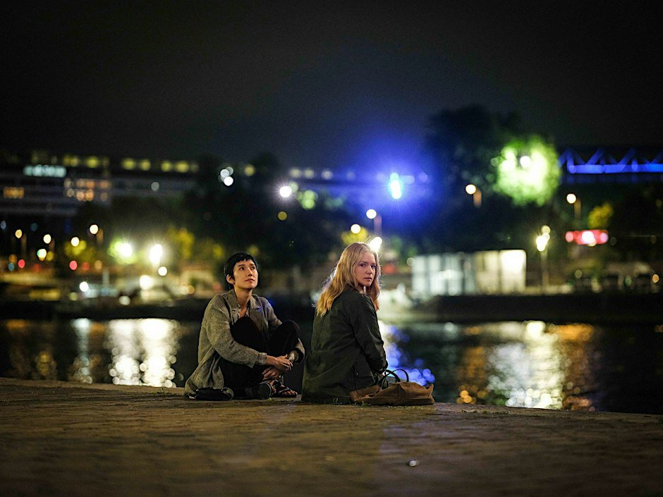 Two women seated on a Paris riverbank at night, city lights reflecting on the water, a still from the film