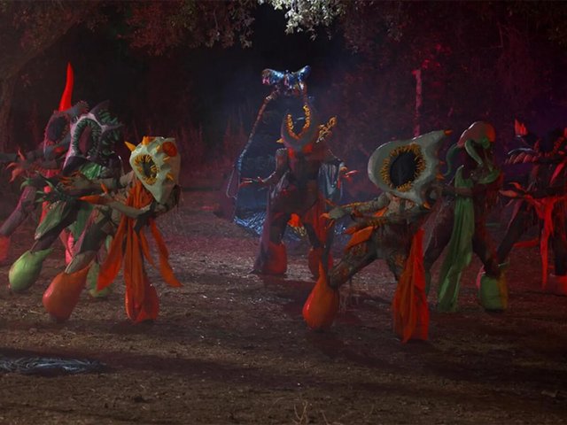 Costumed forest monsters with large mouth-shaped masks performing in a dark, red-lit woodland