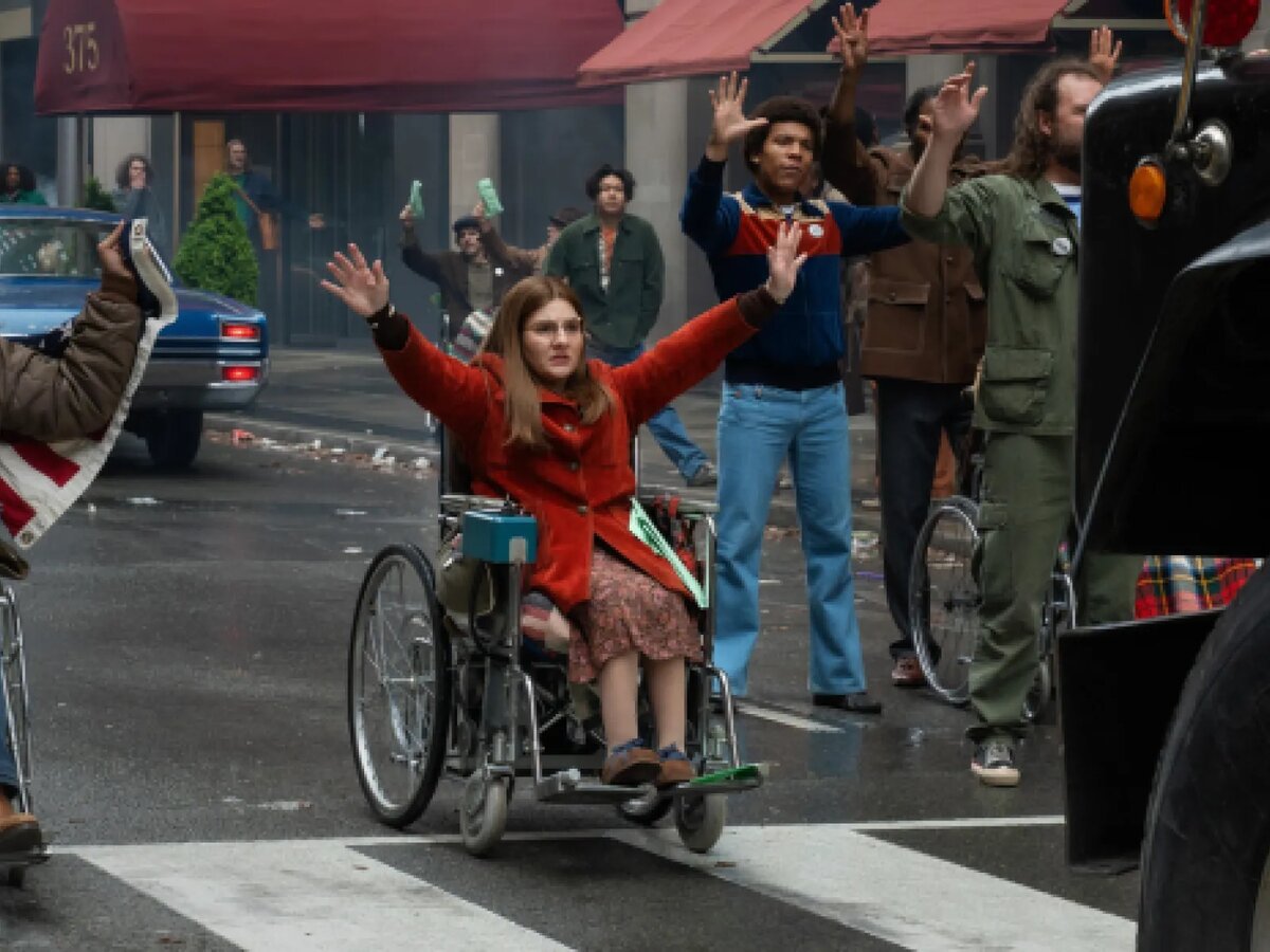 A woman in a red coat raises both arms from a wheelchair in a crosswalk, surrounded by 1970s-dressed protesters