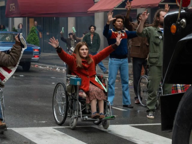 A woman in a red coat raises both arms from a wheelchair in a crosswalk, surrounded by 1970s-dressed protesters