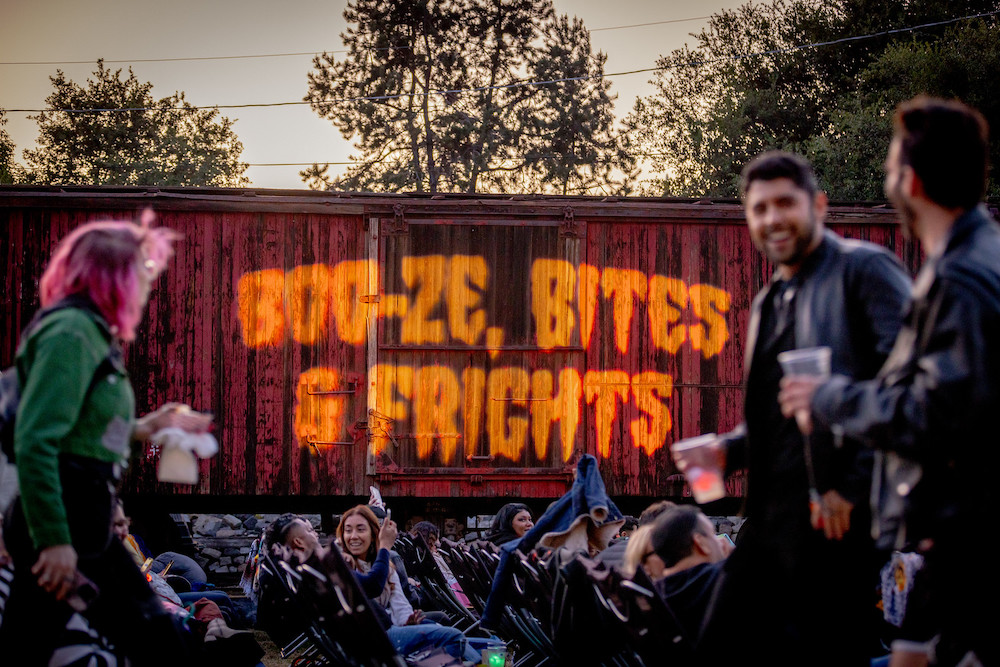 Moviegoers in lawn chairs and a red boxcar with the words Booz-e, Bites and Frights projected on its side at dusk