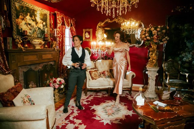 Two performers in Victorian dress standing in a red-walled parlor with chandeliers and a patterned rug