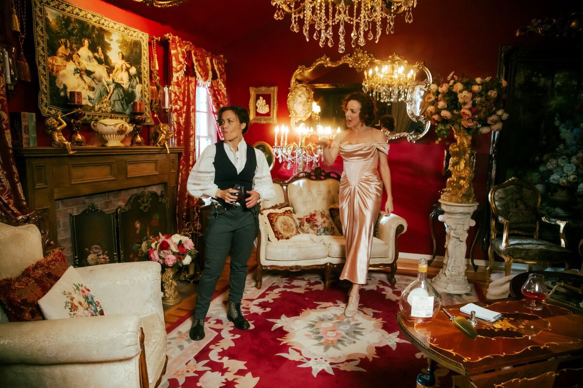 Two performers in Victorian dress standing in a red-walled parlor with chandeliers and a patterned rug