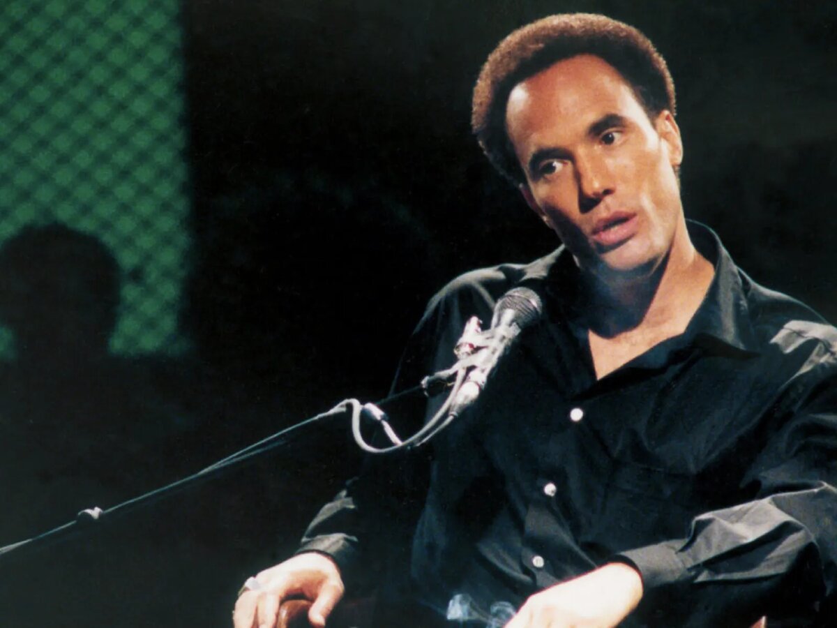 Film still of a man seated at a microphone on a dark stage, from A Huey P. Newton Story