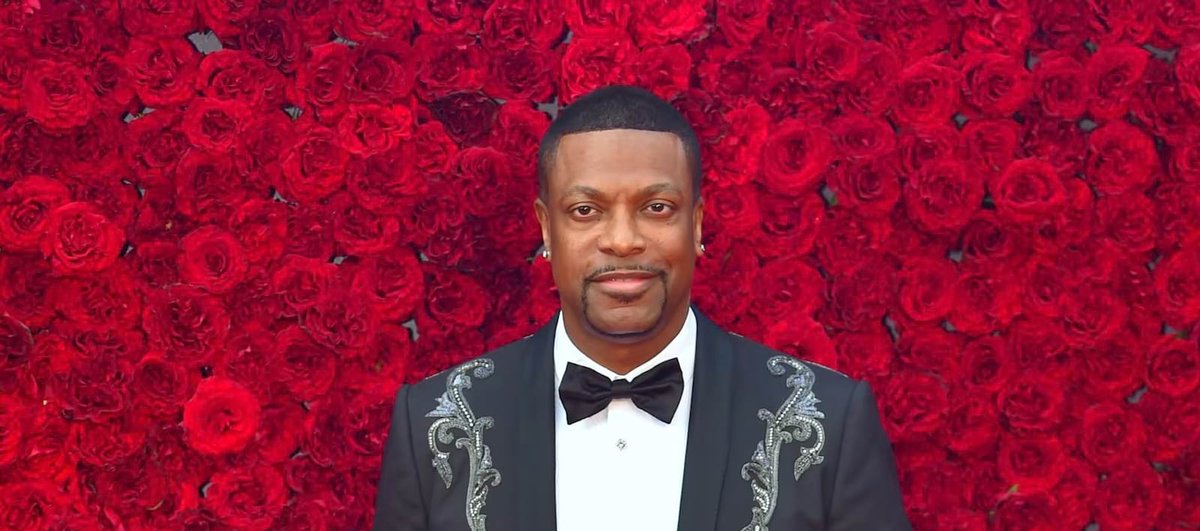 Chris Tucker in a black tuxedo jacket and bow tie in front of a wall of red roses