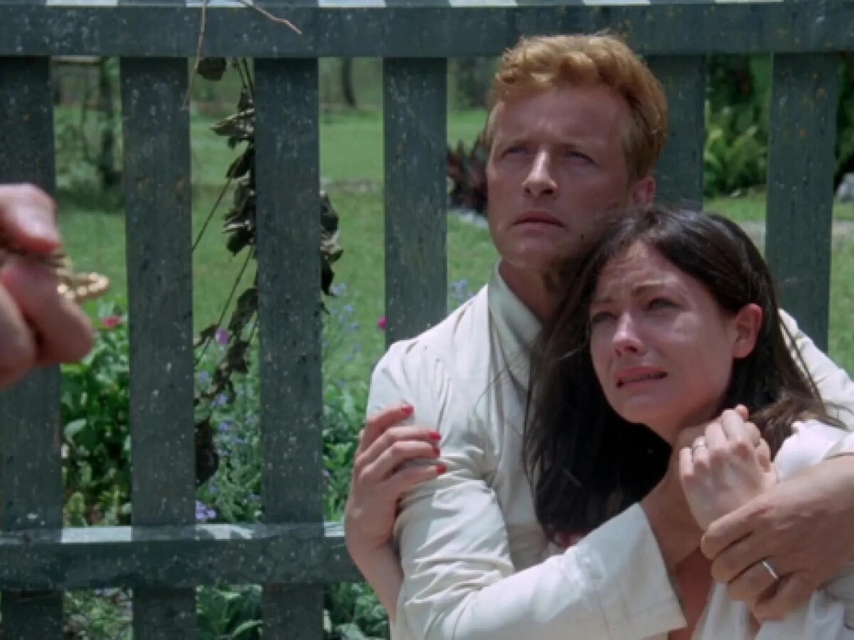 Man and tearful woman clinging to each other in front of a wooden fence in a film still