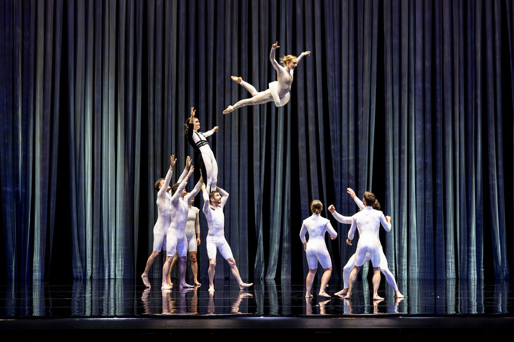 Circa performers in white bodysuits lifting a dancer mid-leap on a stage with blue curtains