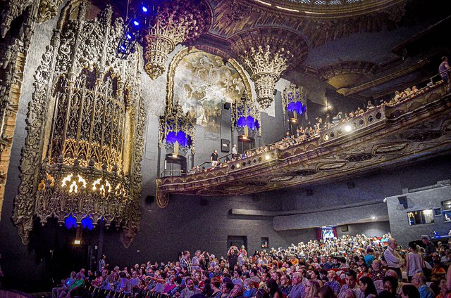 Audience filling the main floor and balcony of a richly ornamented historic theater, lit by blue lanterns