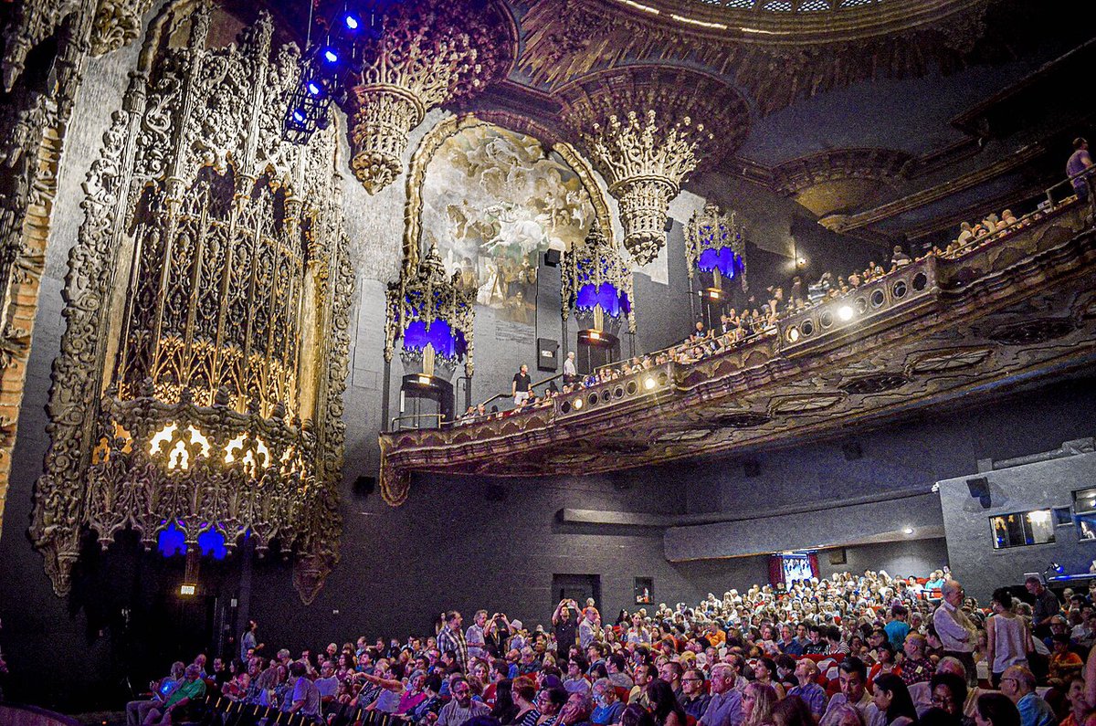 Audience filling the main floor and balcony of a richly ornamented historic theater, lit by blue lanterns