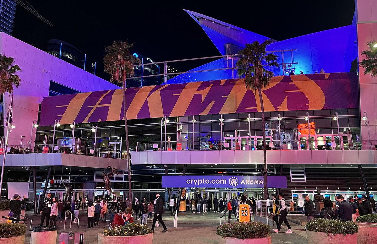 Crypto.com Arena's Star Plaza entrance at night, with crowds walking toward the doors under a purple and gold Lakers banner