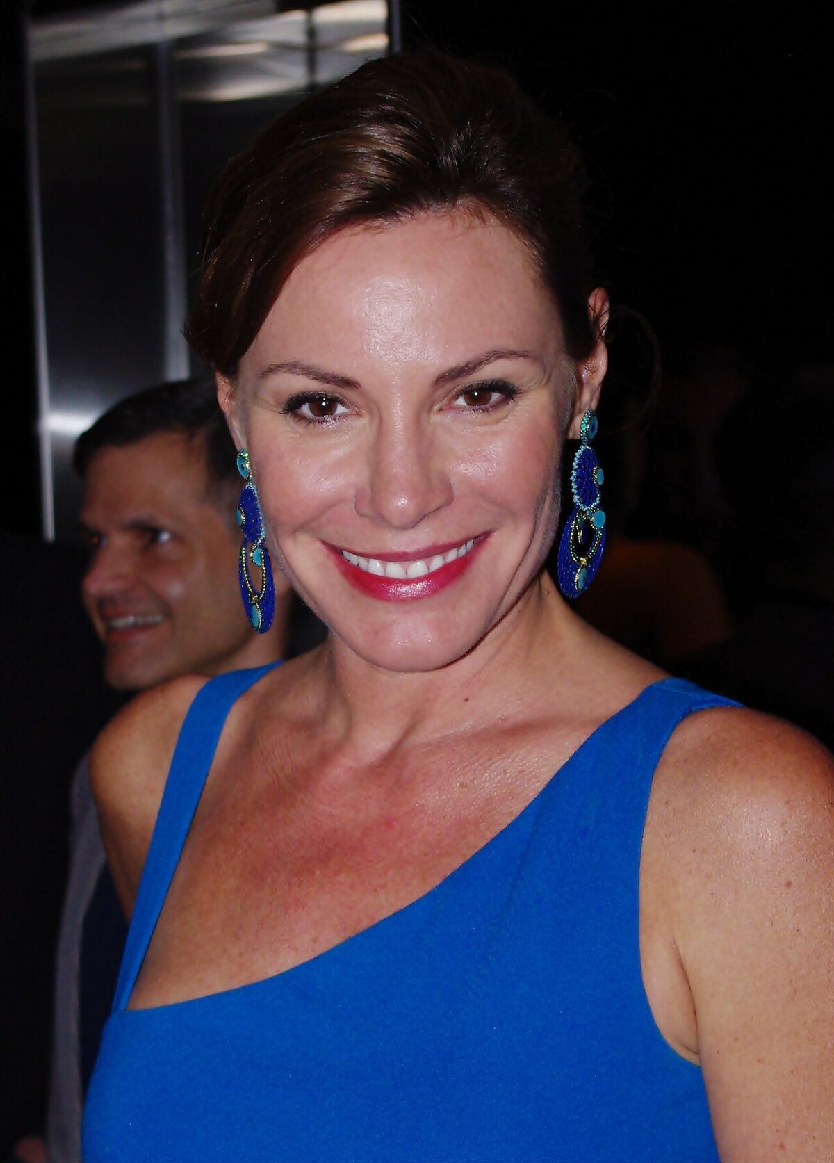 Smiling woman with dark hair pulled back, wearing a bright blue top and large blue beaded earrings