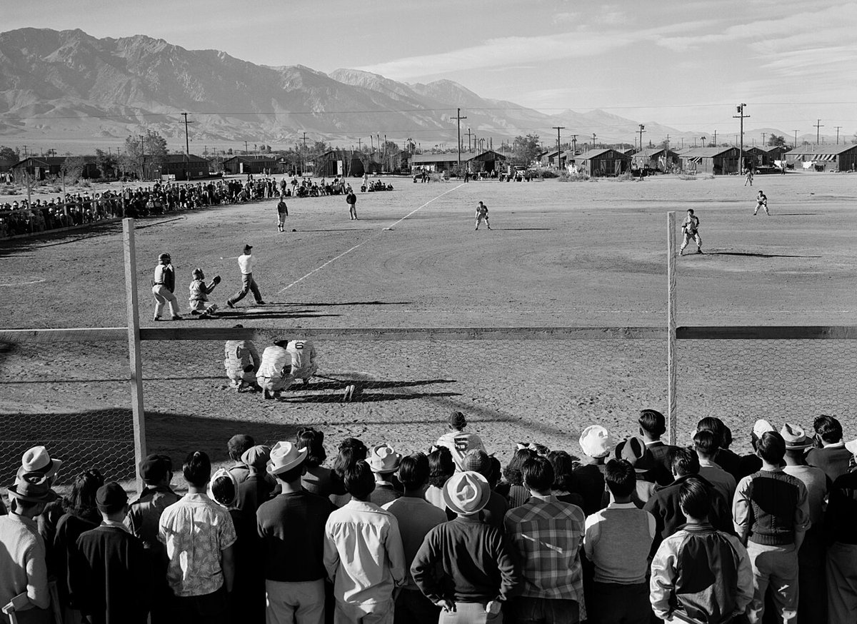 Black and white 1943 photograph by Ansel Adams of Japanese Americans watching a baseball game at Manzanar, with camp buildings and mountains in the background