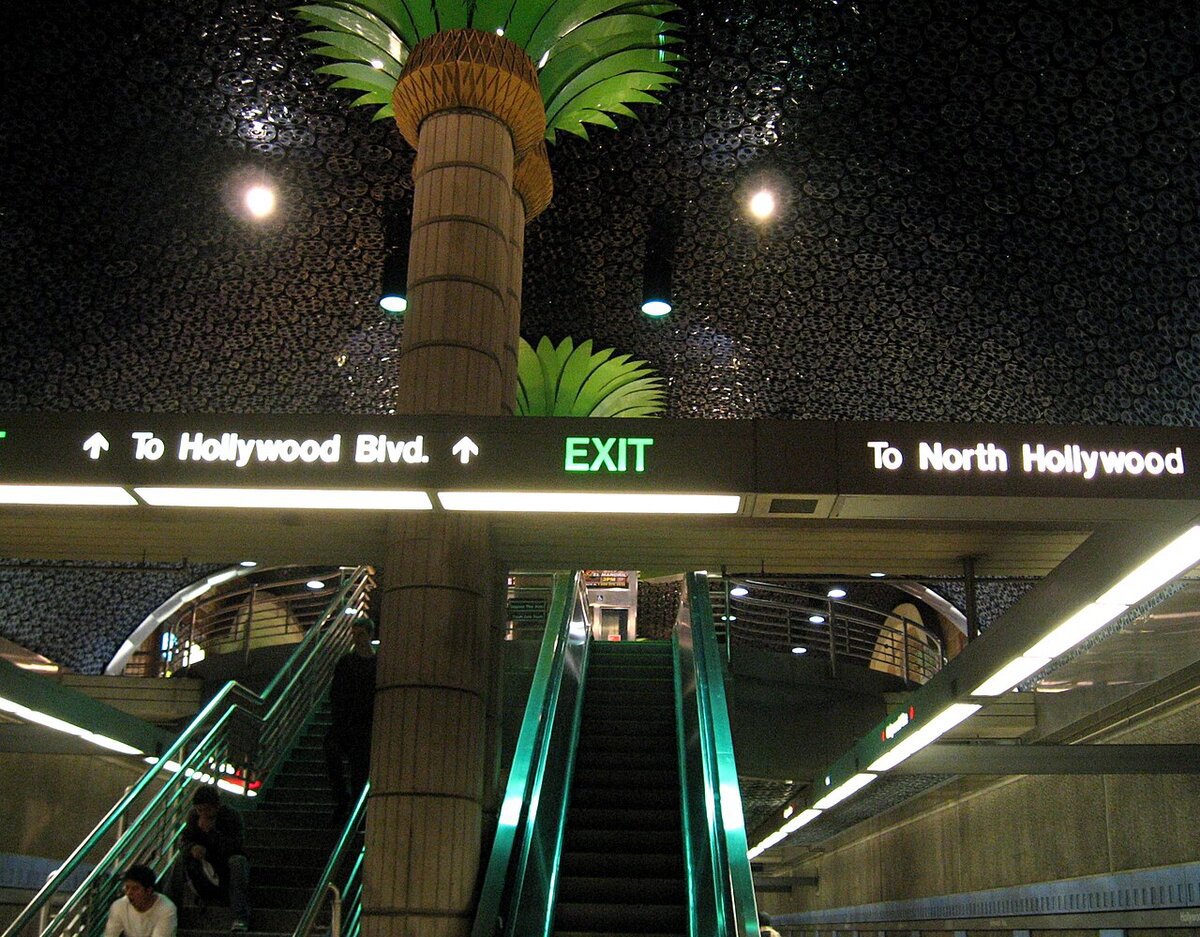 Escalators and a palm tree sculpture inside the Hollywood and Vine Metro station, with signs pointing to Hollywood Blvd.