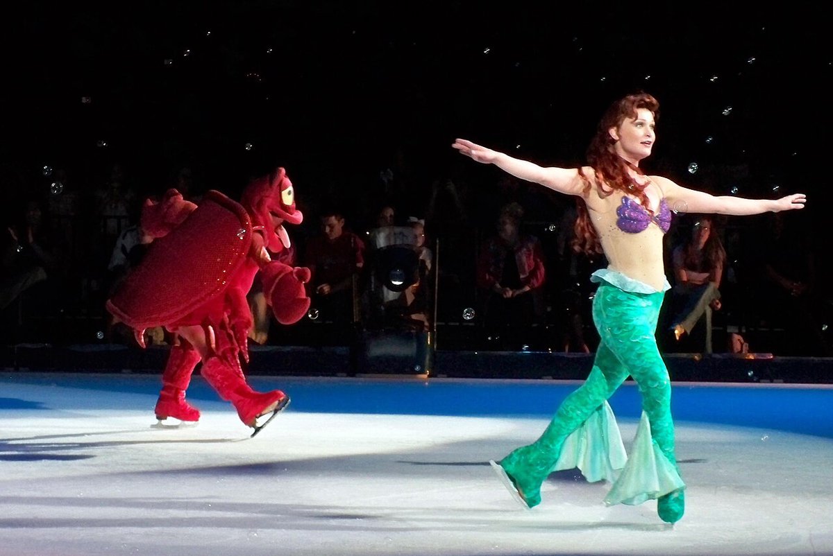 A skater dressed as Ariel glides across a Disney On Ice rink while a skater in a red Sebastian crab costume follows behind
