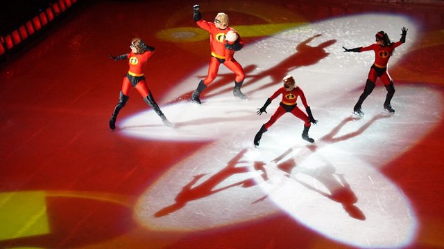 Four skaters dressed as the Incredibles family pose in spotlights on a red-lit Disney On Ice rink