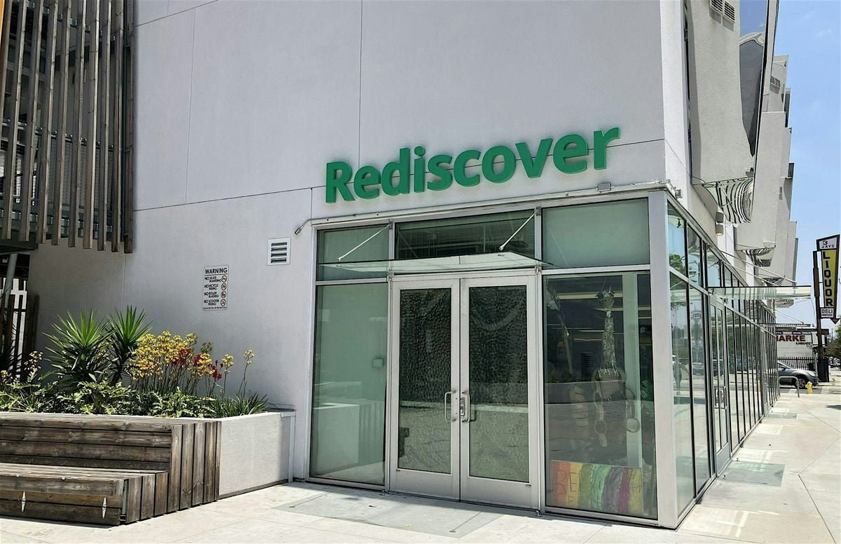 Entrance of the Rediscover Center with its green sign above glass doors on a sunny street corner