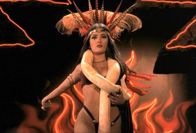 Film still of a woman in a tall feathered headdress with a pale snake draped over her shoulders against neon flames