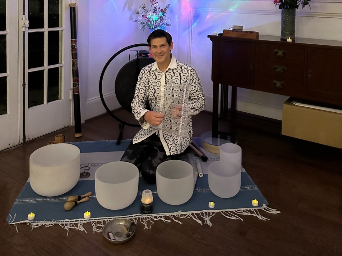 Sound healer seated on a woven rug among frosted crystal singing bowls, a gong and small candles in a softly lit living room