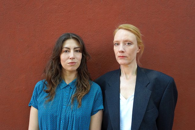 Two women standing side by side against a rust-red wall, one in a blue patterned shirt and one in a dark blazer