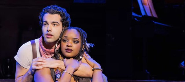 Two performers in the Hadestown tour embracing on a dimly lit stage, one in a pink bandana