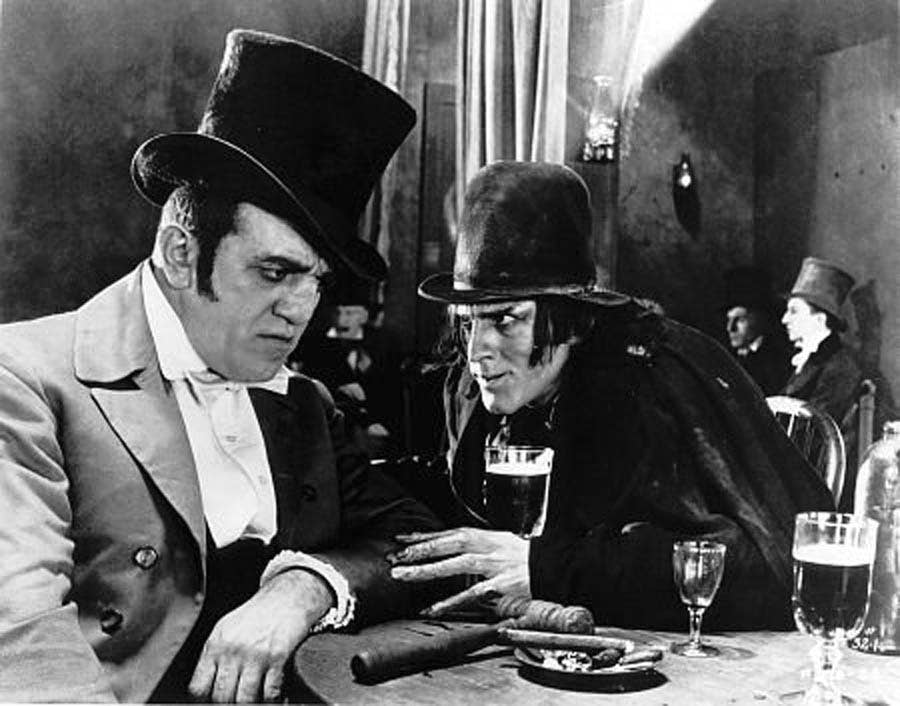 Black and white film still of two men in top hats leaning toward each other across a tavern table