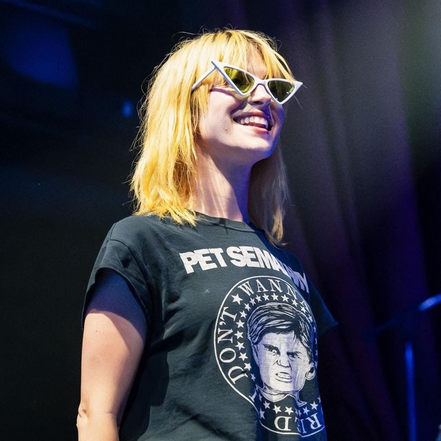 Hayley Williams smiling on stage in white cat-eye sunglasses and a black band T-shirt under blue stage light