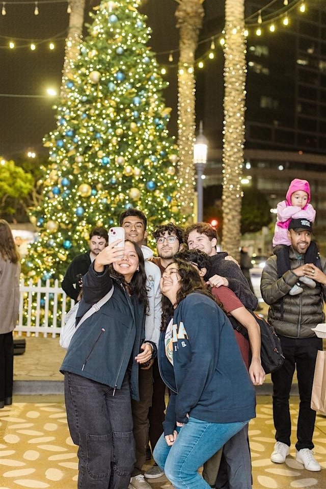 Friends take a selfie in front of a large lit holiday tree with gold and blue ornaments, palm trees wrapped in string lights behind them