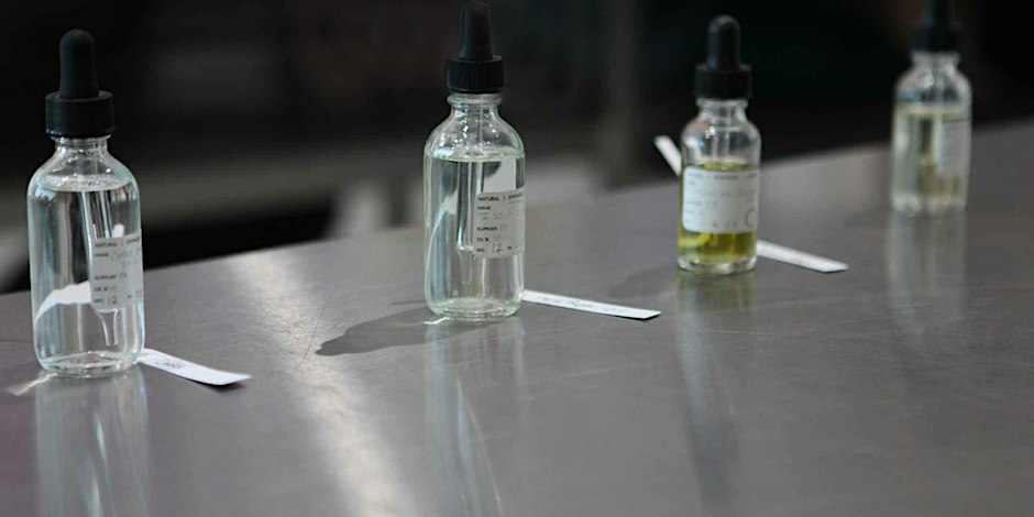 Labeled glass dropper bottles of perfume materials lined up with paper scent strips on a steel lab table