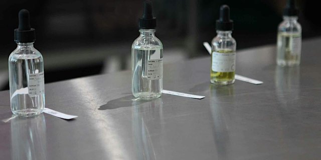 Labeled glass dropper bottles of perfume materials lined up with paper scent strips on a steel lab table