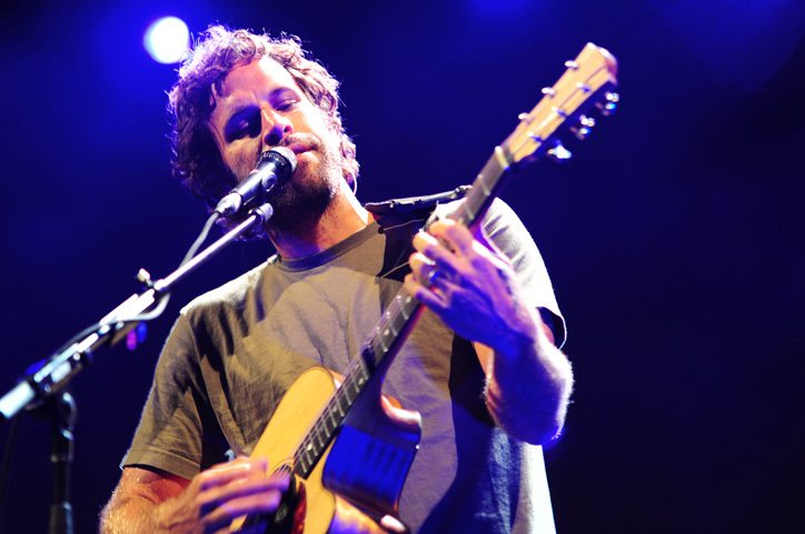 Jack Johnson singing into a microphone and playing acoustic guitar under blue stage lights