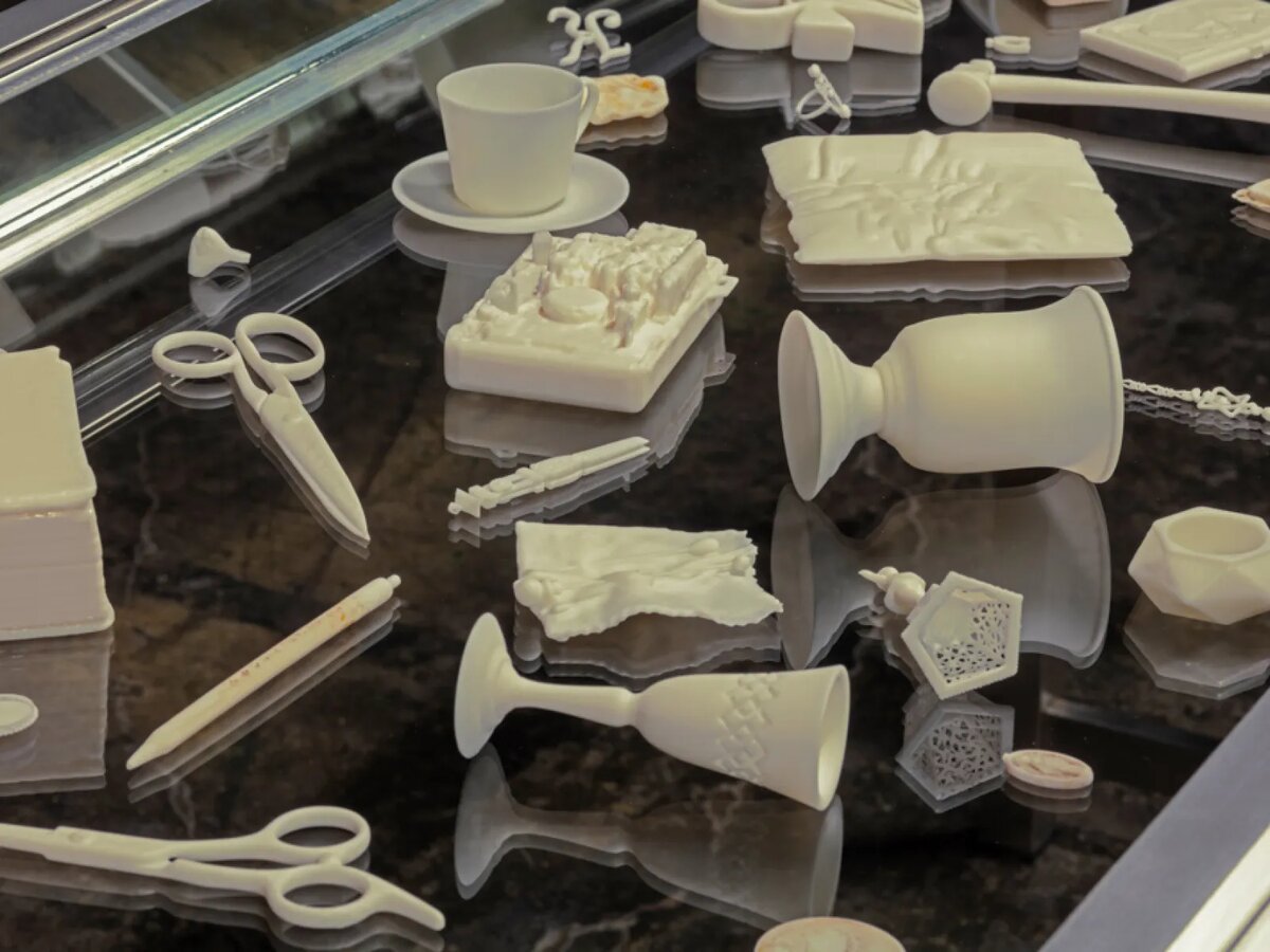 White 3D-printed replicas of keepsakes, including scissors, goblets and a teacup, arranged on a dark glass surface