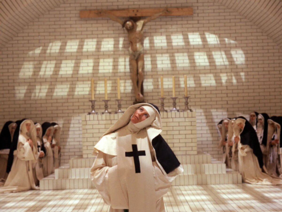 A nun in a white habit with a black cross looks upward before a crucifix, with rows of kneeling nuns in a white-tiled chapel