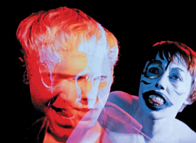 Double-exposed film still of a red-lit man's face beside a figure in blue and white face paint with a wide open mouth