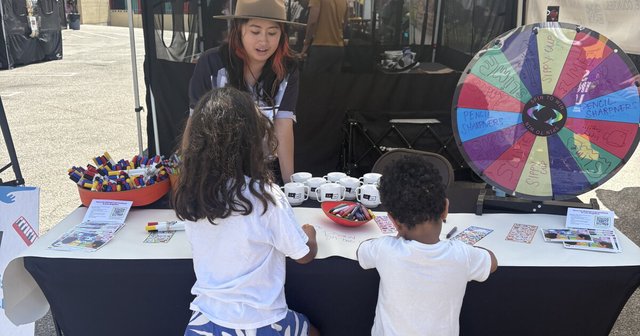 Two children drawing at an LAist table with a prize wheel and markers while a staffer in a hat talks with them