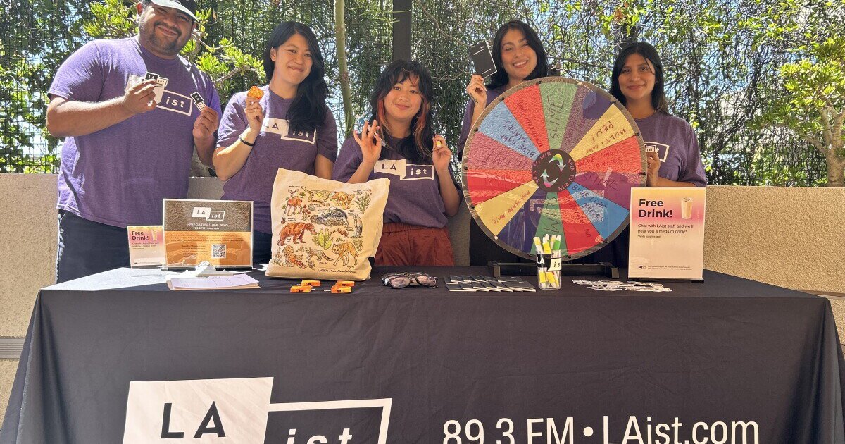Five LAist staffers in purple shirts smile behind a table with a prize wheel, tote bag and free drink signs