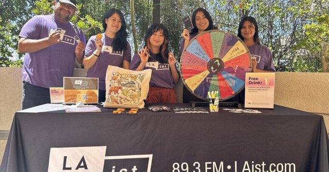 Five LAist staffers in purple shirts smile behind a table with a prize wheel, tote bag and free drink signs