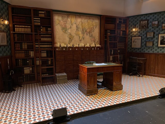 An empty theatre set of a book-lined study with a wooden desk on a black-and-white tiled floor, lit with no actors on stage