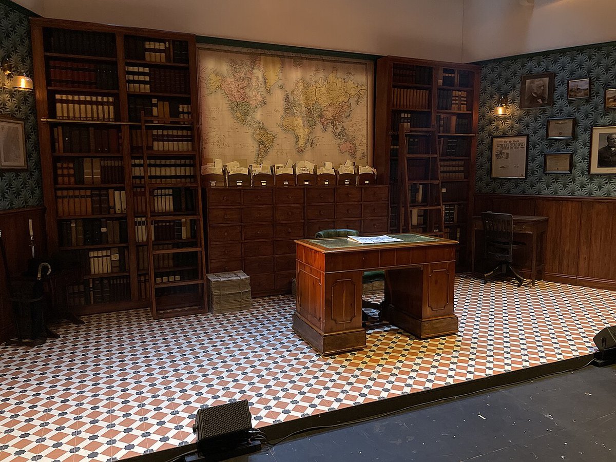 An empty theatre set of a book-lined study with a wooden desk on a black-and-white tiled floor, lit with no actors on stage