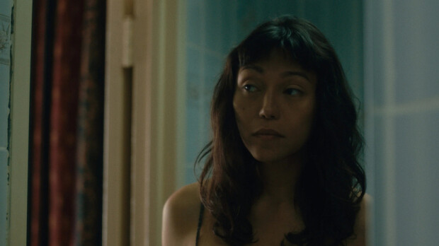 A still from Lingua Franca showing a woman with dark hair and bangs glancing sideways in a dim room with pale teal walls