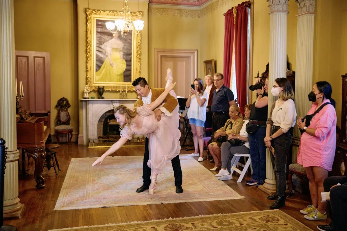 A ballerina in a pink dress dips backward with her partner in a Victorian parlor at Heritage Square Museum as guests watch from the edges of the room