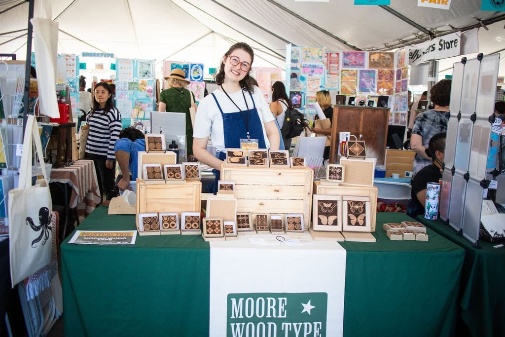 Vendor smiling behind a table of wood-type printed coasters and prints at a crowded tented printing fair