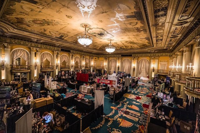 Vendor tables and book displays filling a gilded ballroom with a painted ceiling and chandeliers