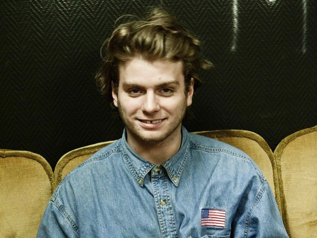 Mac DeMarco smiling in a denim button-down shirt, seated on a gold velvet theater seat against a dark wall