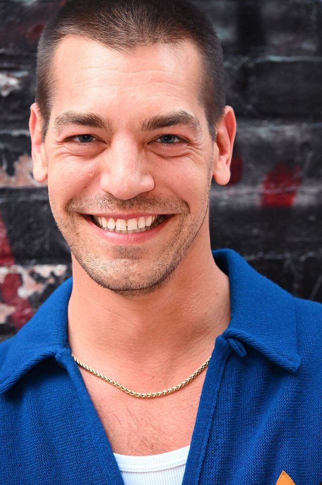 Close-up portrait of a smiling man with short dark hair, stubble and a blue knit collared shirt in front of a dark brick wall