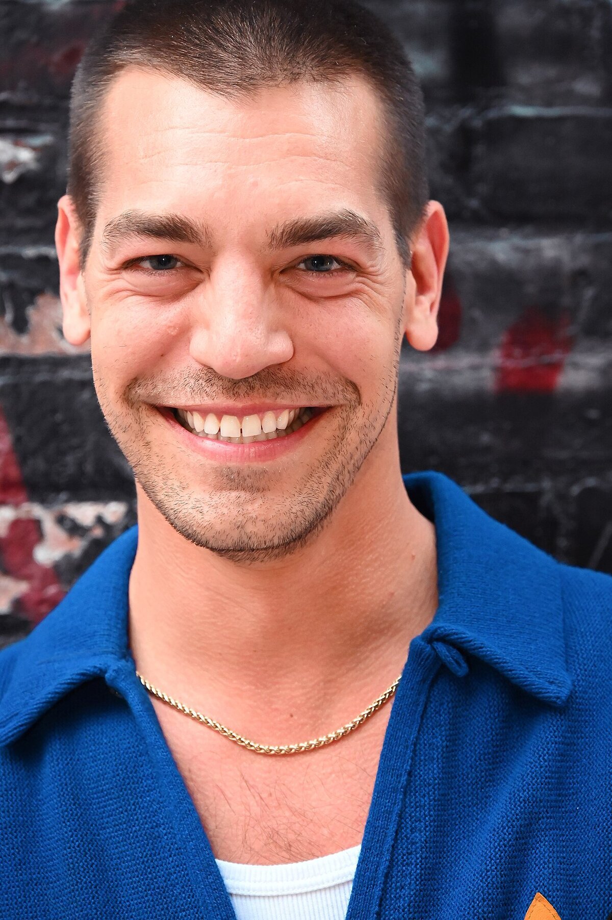 Close-up portrait of a smiling man with short dark hair, stubble and a blue knit collared shirt in front of a dark brick wall