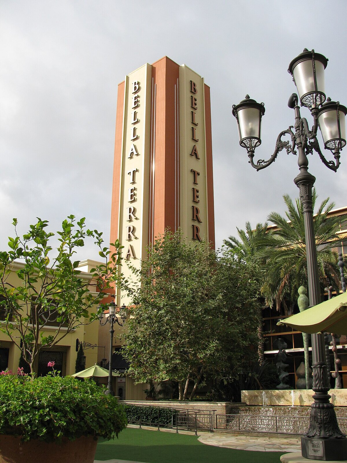 Tall Bella Terra tower with vertical lettering, framed by trees, palms and ornate lampposts under a cloudy sky