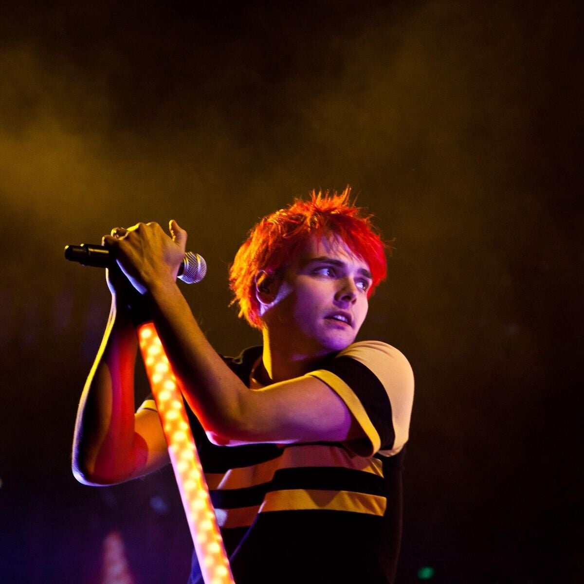 Singer with bright red hair gripping a microphone stand lit with warm stage lights in a haze of smoke