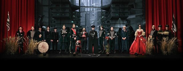 Promotional artwork of band members in black military-style uniforms standing in a row before red stage curtains